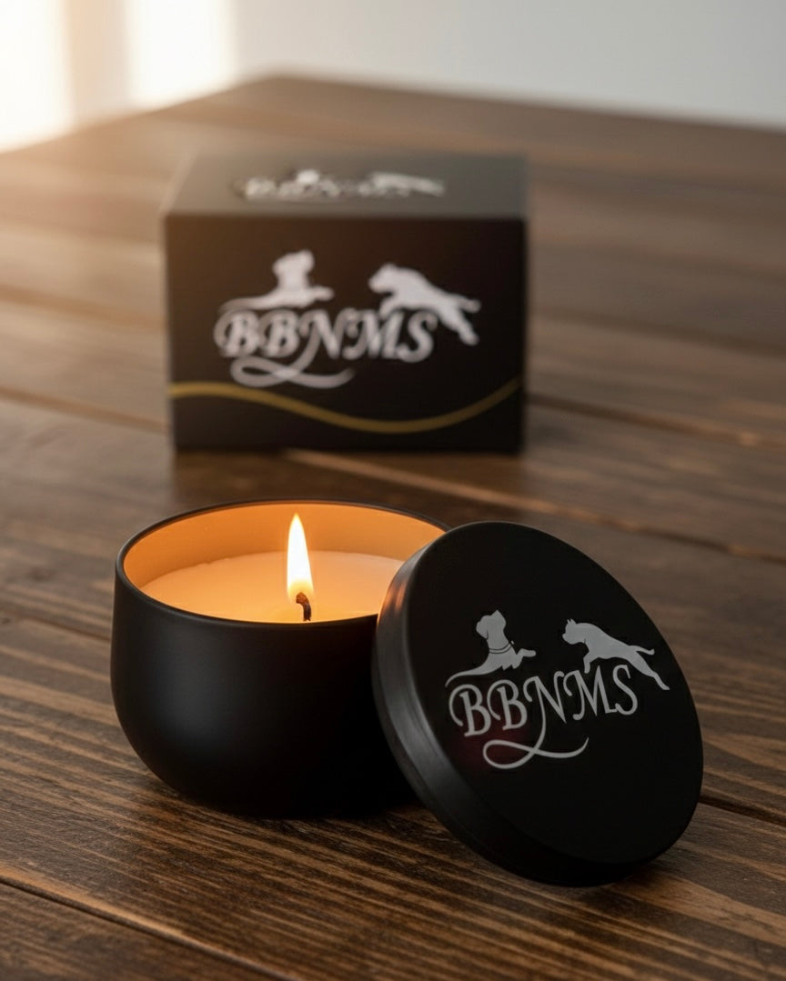 Black candle tin, lit candle and lid featuring 'BBNMS' logo on a wooden surface. The products box is softly blurred behind it.